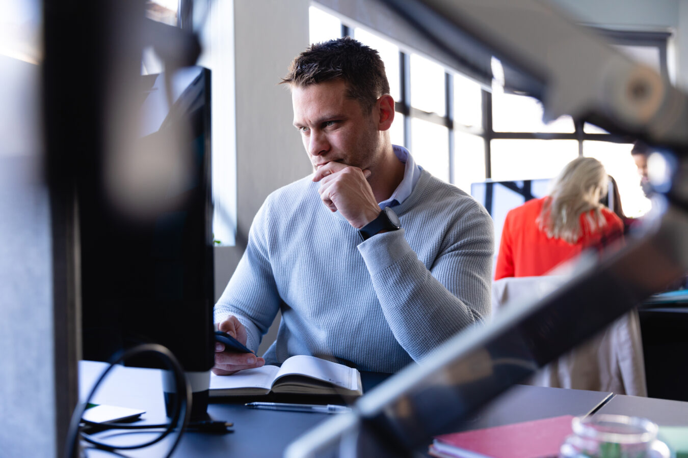 Front view of a Caucasian man wearing smart clothes, working in a modern office, using a desktop computer and smartphone, with other colleagues working in the background.